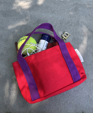 Wear One's At Studio Tote with purple handles, shown outdoors on a gray concrete surface with various items like a tennis ball, earplugs, and a small bottle peeking out of the top.