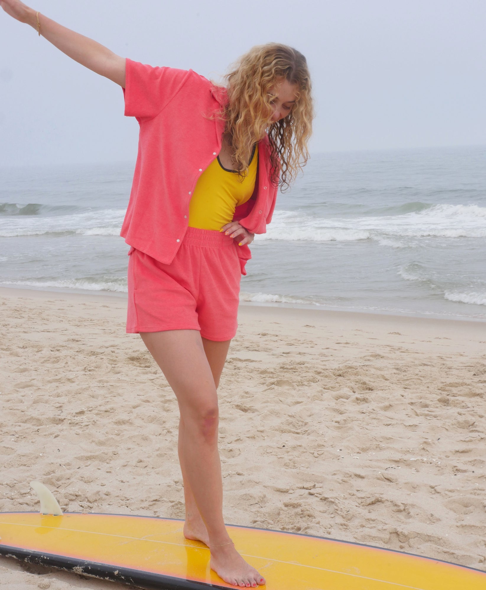 Wear One's At Aqualina Terry Button Down in Aperol Pink on Model Standing on a Surfboard Side View